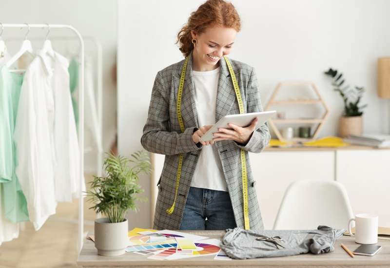 Cheerful Red-Haired Female Fashion Designer Using Tablet Computer Browsing Internet In Dressmaking Studio. Copy Space